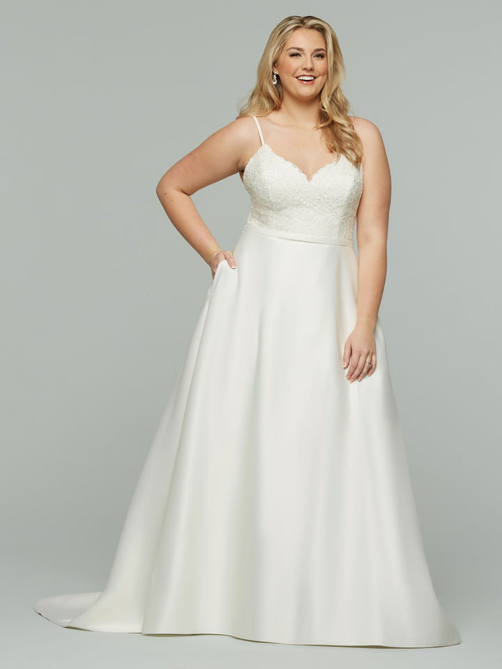 A woman smiles with one hand in her pocket, wearing the Avery Austin "Oakley" wedding dress in ivory—a satin A-line gown with a lace bodice and flowing skirt—posed against a plain light gray background.