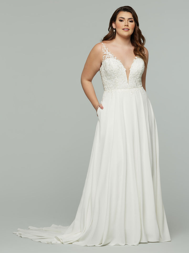 A woman stands against a gray background wearing the Skye by Avery Austin: an ivory A-line wedding gown with pockets, a sleeveless lace bodice, deep V neckline, and flowing chiffon skirt.