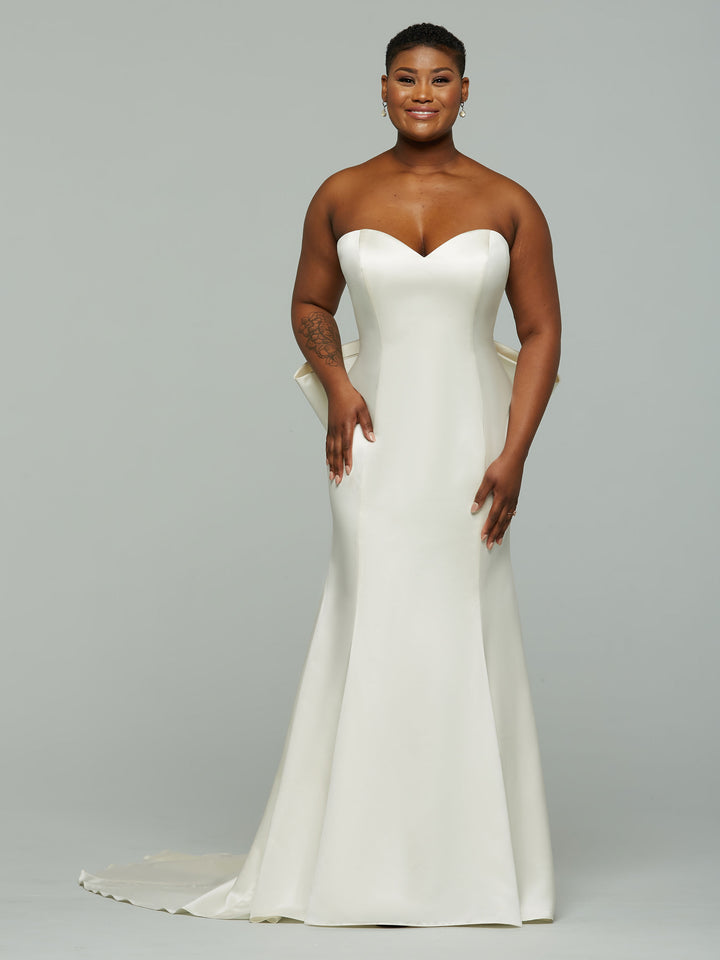 A woman smiles in the Avery Austin TH-Tori ivory satin mermaid wedding dress with a strapless sweetheart neckline, long train, and detachable satin bow, standing against a plain light gray background.