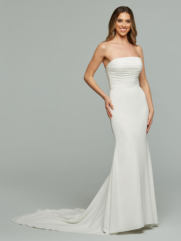 A woman stands smiling against a plain light gray background, wearing the Avery Austin "Vera" wedding dress in ivory, featuring a modern strapless design with a ruched bodice and flowing train.