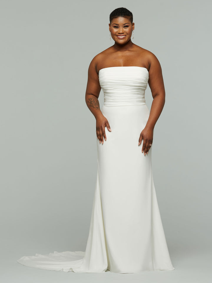 A woman with short hair smiles in the Avery Austin TH-Vera wedding dress, a modern ivory strapless gown with a fitted bodice and slight train, standing against a plain light gray background.