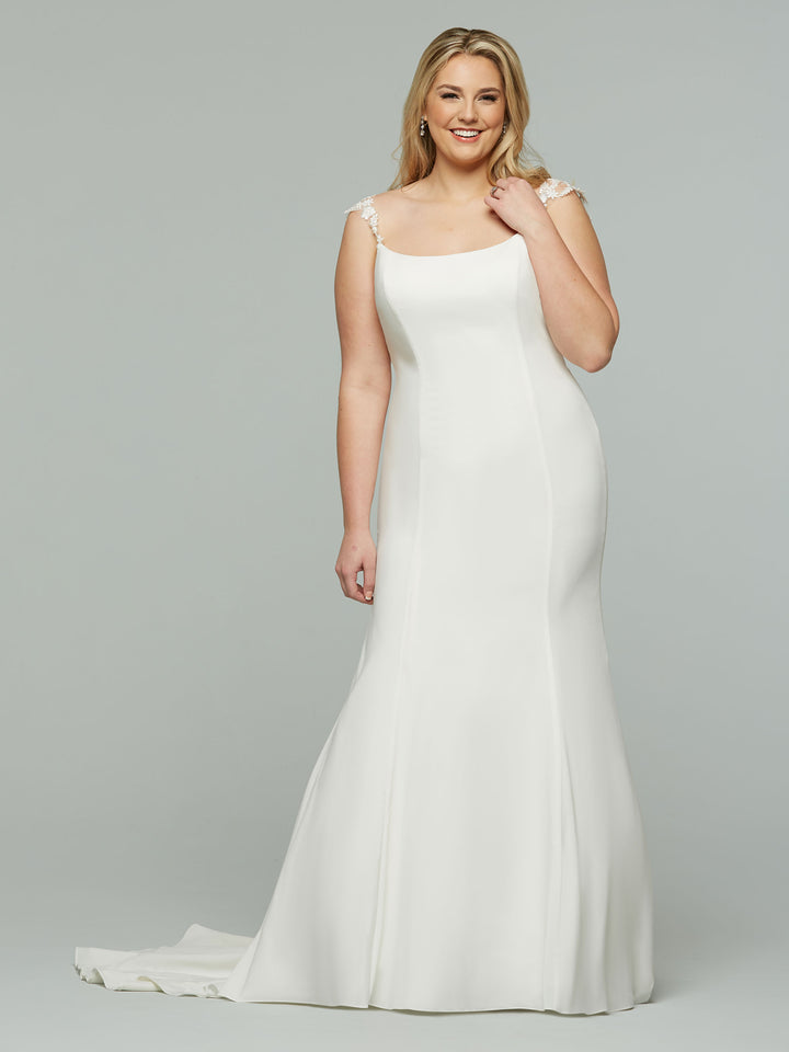 A woman smiles in front of a gray backdrop, holding one shoulder strap while wearing the Avery Austin Whitley—an ivory, floor-length wedding dress with a fitted bodice, cap sleeves, and a slight train.