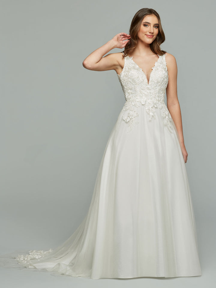 A woman smiles and touches her hair while wearing the ivory Aspen wedding dress by Avery Austin, featuring a sleeveless A-line silhouette with lace bodice detailing and a flowing tulle skirt, set against a light gray background.