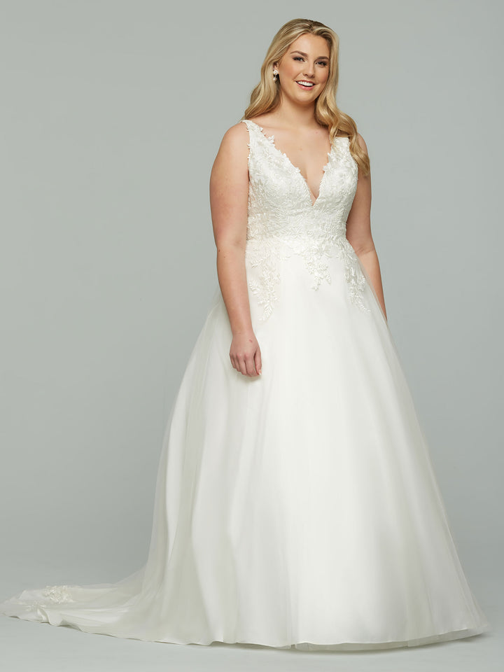 A woman in the Aspen ivory wedding dress by Avery Austin, featuring a sleeveless V-neck, lace bodice details, and a flowing A-line skirt, stands against a plain light gray background, smiling softly.