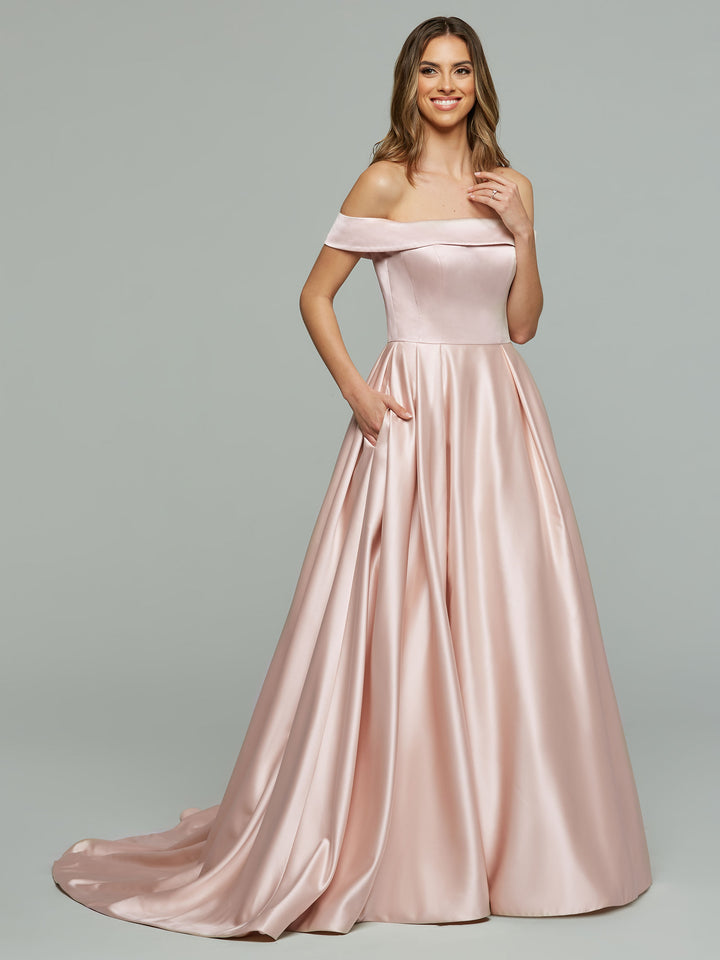A woman smiles with one hand in her hair and the other in her pocket, modeling the Avery Austin Blake wedding dress in blush—a fitted bodice and full satin skirt, off-the-shoulder style—against a plain gray background.