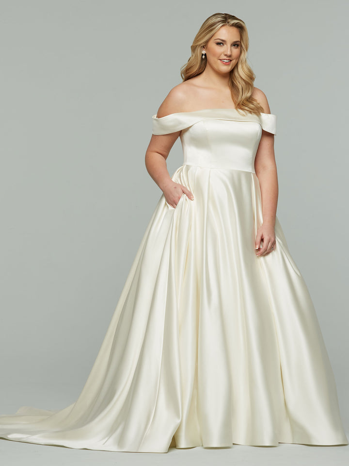A woman wearing Avery Austin’s Blake blush off-the-shoulder satin ball gown wedding dress, featuring a fitted bodice and full skirt, poses against a light gray background with long blonde hair, smiling and one hand in her pocket.