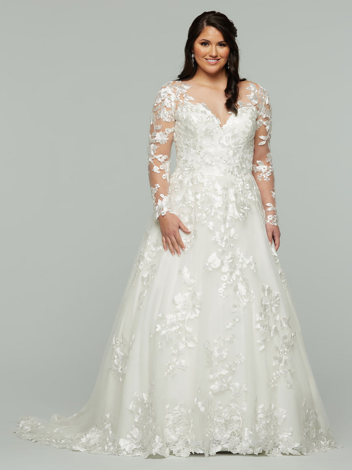 A woman smiles against a light gray background wearing the Avery Austin Dylan wedding dress in ivory, featuring sheer long sleeves, floral appliqué details, an illusion design, and a fitted bodice.