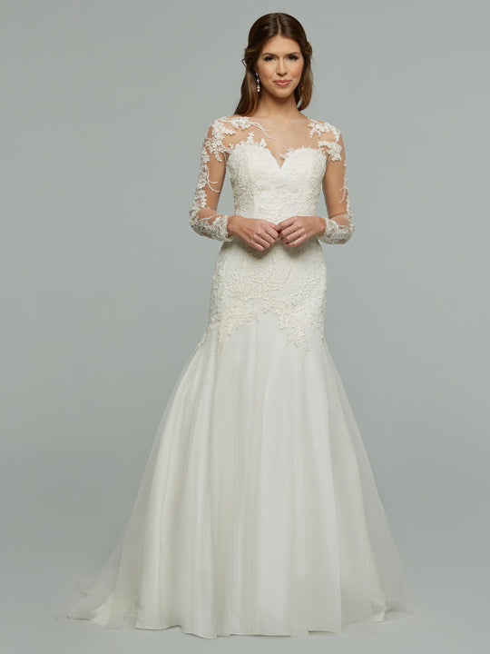 A woman wears the Avery Austin "Hallie" wedding dress: a white, floor-length lace gown featuring long sheer sleeves, a fitted bodice, and a flowing skirt against a plain light gray background.