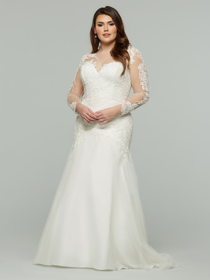 A woman wearing the Avery Austin Hallie, a white lace fit-and-flare wedding dress with long sleeves and a flowing skirt, stands against a plain gray background, looking confidently at the camera.