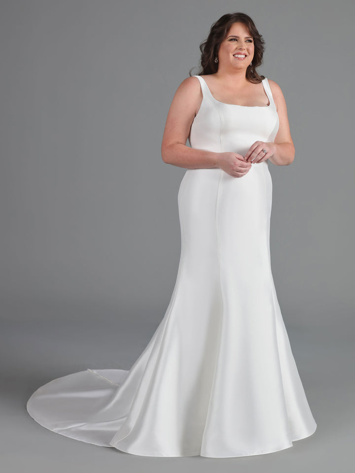 A woman wearing the Avery Austin Dakota mermaid wedding dress in ivory, featuring a sleeveless fitted bodice and flared train, stands smiling against a plain gray background.