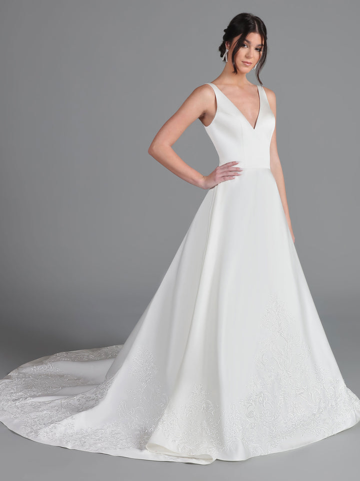 A woman models the Avery Austin "Sierra" gown in ivory, featuring a sleeveless v-neck and a long embroidered train, standing before a plain gray background with one hand on her hip.