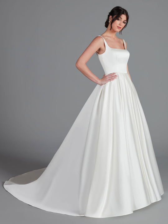 A woman stands in a studio wearing the Avery Austin "Chelsea" wedding dress in ivory, featuring a classic square neckline, fitted bodice, and a full-length skirt with a short train. The background is plain gray.