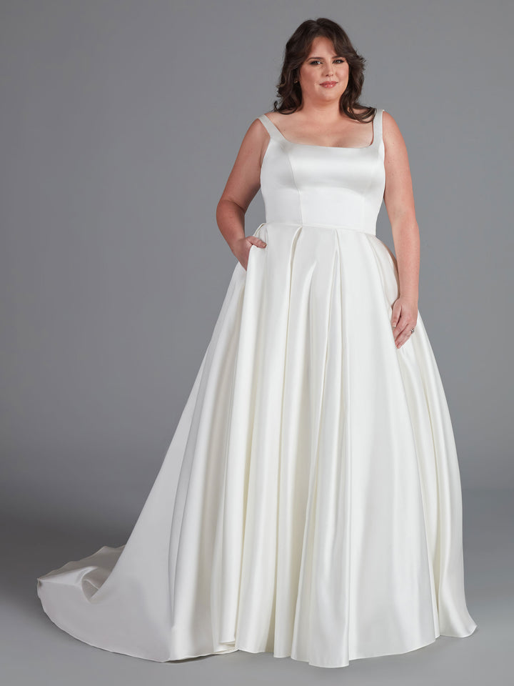 A woman in the Avery Austin "Chelsea" gown—a simple ivory satin ball gown with a fitted bodice and full, pleated skirt—smiles gently against a gray background, one hand tucked in her pocket.