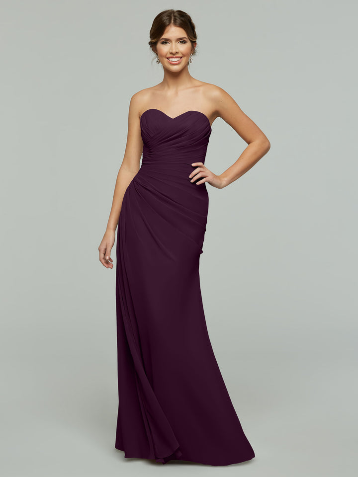 A woman wearing the Avery Austin TH-80028 aubergine strapless chiffon gown with a sweetheart neckline stands smiling, one hand on her hip, against a plain light gray background.