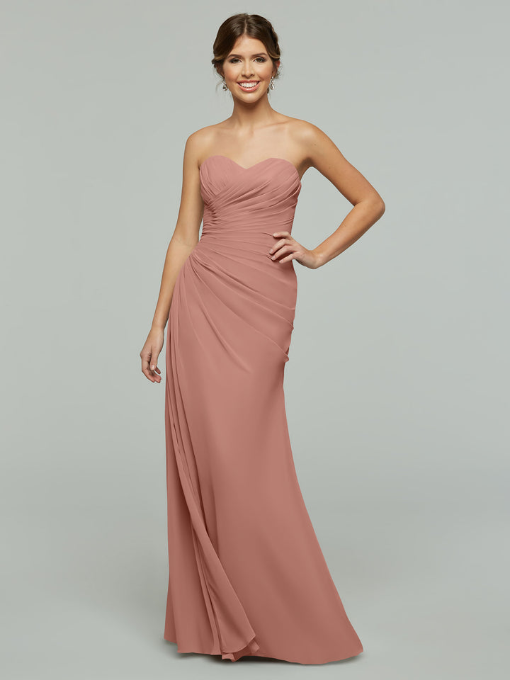 A woman stands against a plain gray background wearing Avery Austin’s 80028, a strapless floor-length chiffon dress in dusty rose with ruched detailing and a subtle sweetheart neckline.