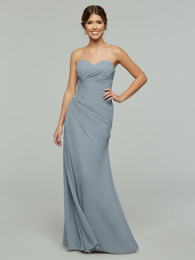 A woman in a Dusty Blue Avery Austin 80028 floor-length chiffon dress with a ruched bodice, sweetheart neckline, and draped skirt stands against a plain light gray background.