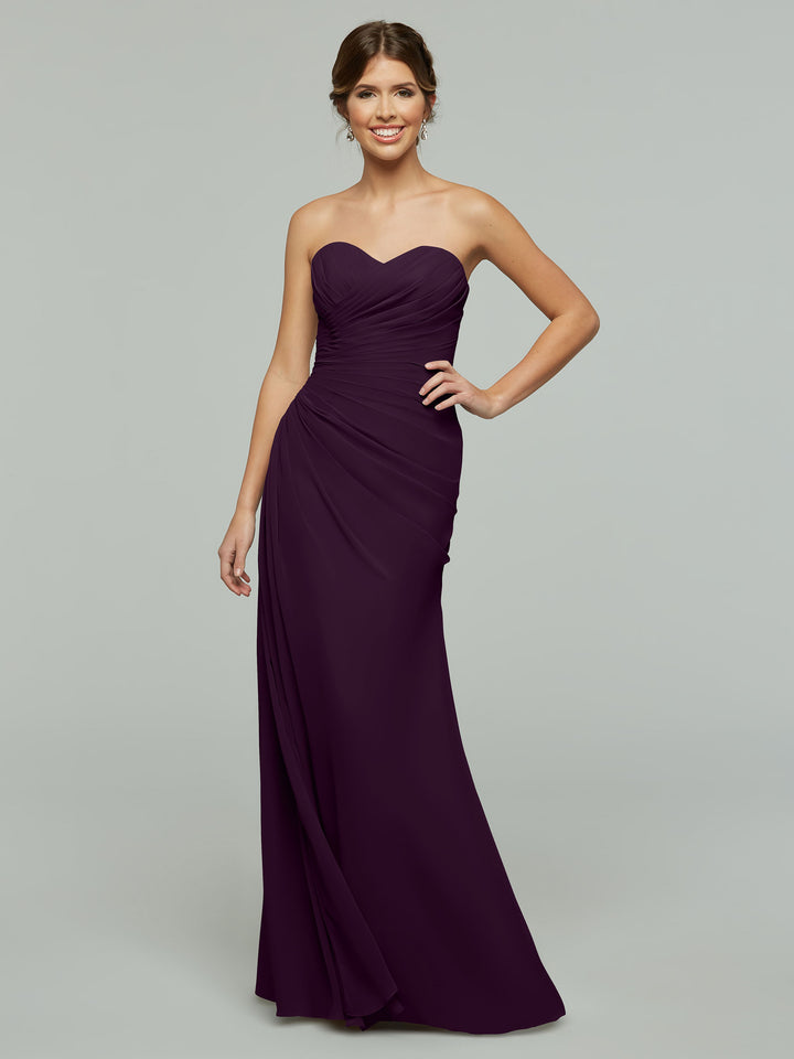 Model wearing Avery Austin’s strapless, floor-length chiffon dress (Style 80028) in dark purple, featuring a ruched bodice and subtle sweetheart neckline, smiling against a plain gray background.