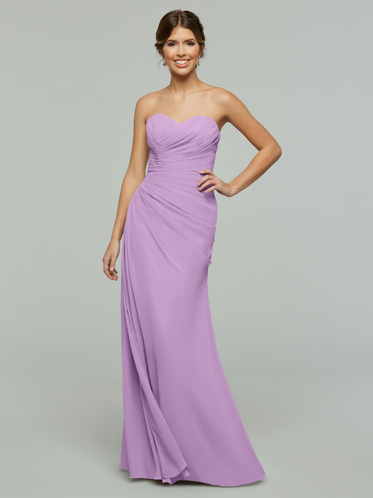 A woman smiles while wearing Avery Austin’s 80028, a lilac chiffon gown featuring a ruched bodice, sweetheart neckline, and draped, floor-length skirt. The elegant dress stands out against a plain light gray background.