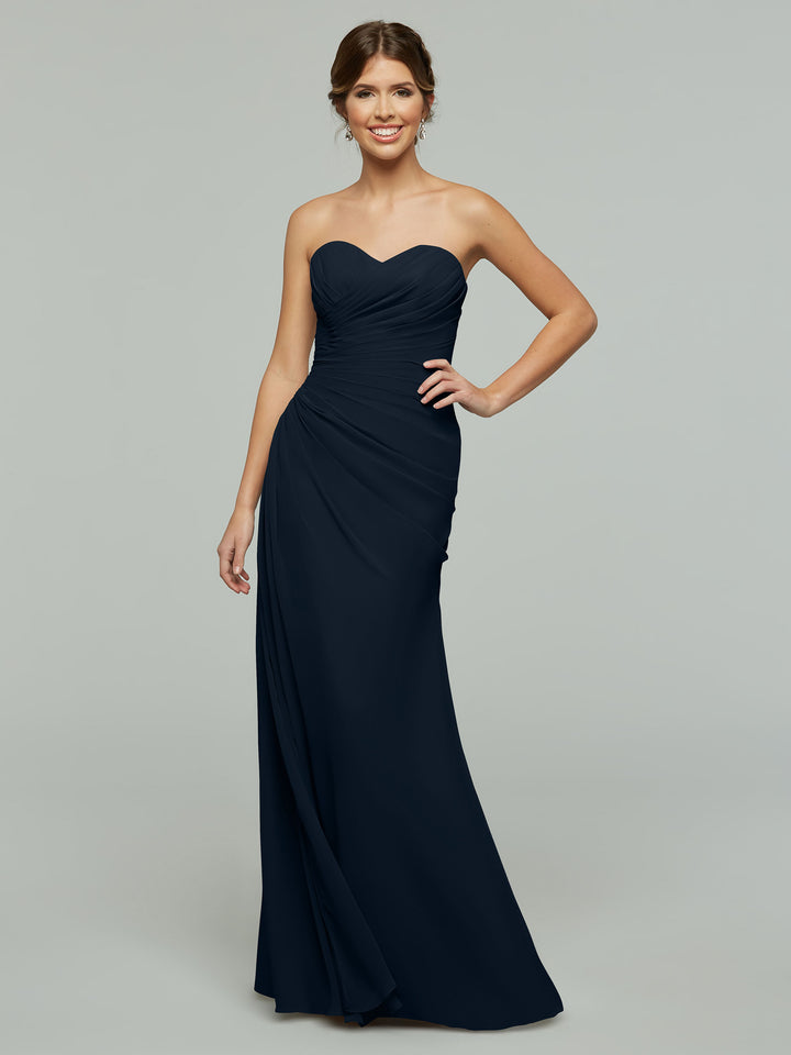 A woman in the Avery Austin 80028 dress, a midnight-colored, strapless chiffon gown with a sweetheart neckline, stands smiling with one hand on her hip against a plain gray background.