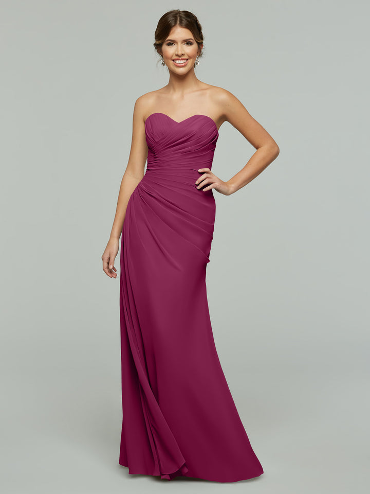 A woman wearing Avery Austin’s strapless, floor-length chiffon dress (style 80028) with a sweetheart neckline stands against a grey background, smiling with one hand on her hip.