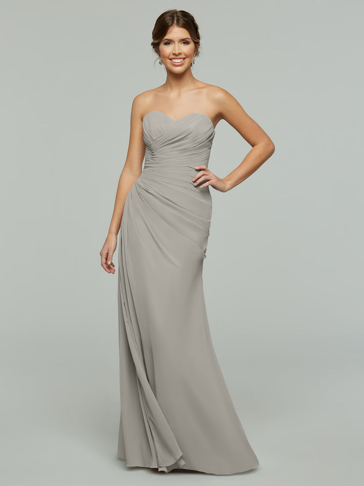 A woman smiles while wearing Avery Austin’s 80028, a strapless floor-length chiffon dress featuring ruched details and a subtle sweetheart neckline, set against a plain light background.