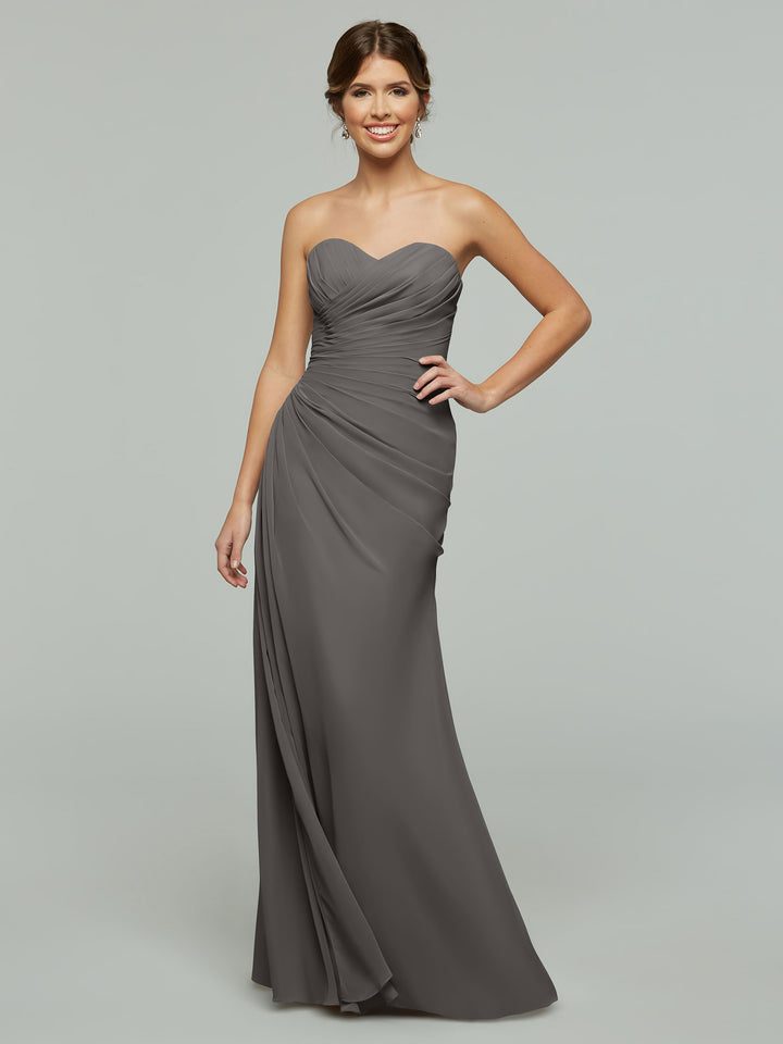 A woman wearing Avery Austin’s 80028 floor-length dress with a sweetheart neckline smiles with one hand on her hip against a plain light gray background.