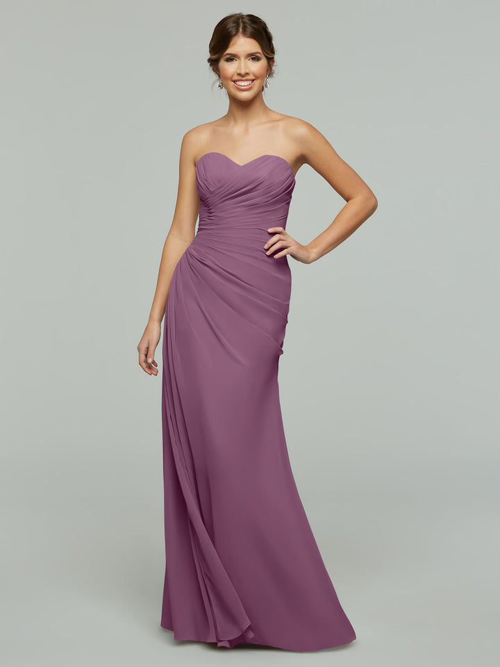 A woman wears Avery Austin’s floor-length mauve chiffon dress (Style 80028) with a sweetheart neckline and ruched detailing, standing against a plain light gray background.