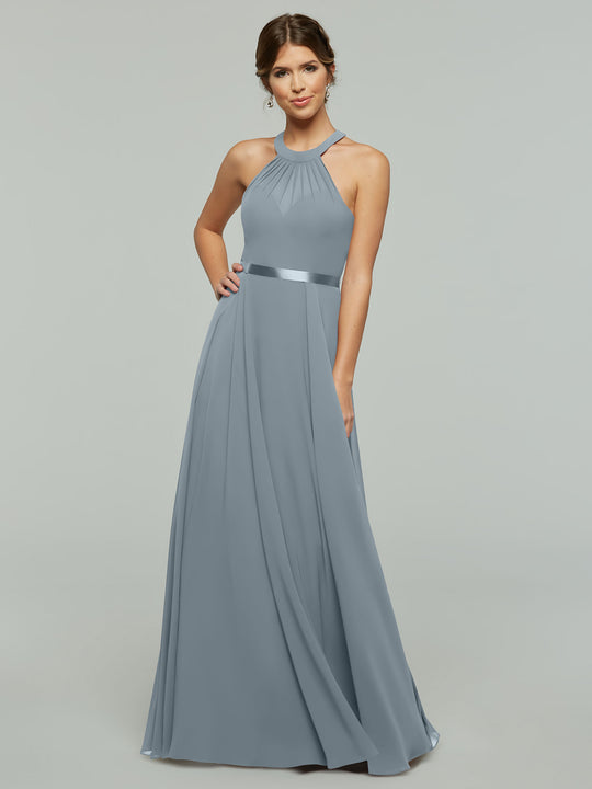 A woman poses against a plain light gray background wearing Avery Austin’s 80038—a dusty blue, floor-length, sleeveless chiffon A-line dress with a halter neckline and satin waistband.