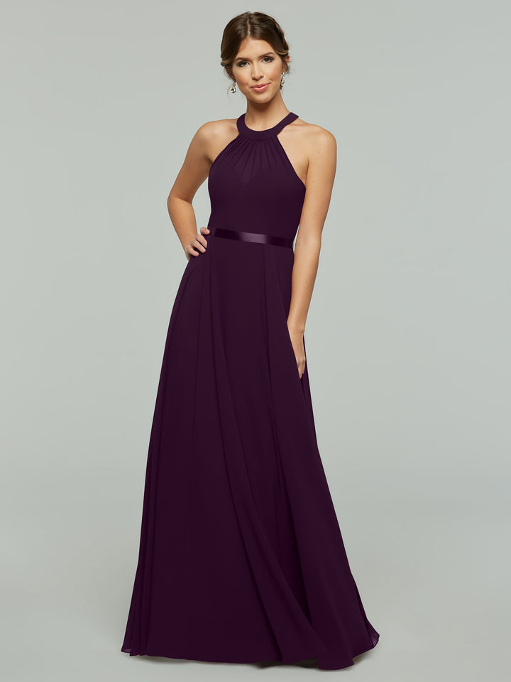 A woman in Avery Austin’s 80038, a floor-length sleeveless chiffon A-line dress with a halter neckline and pleated skirt, stands smiling with one hand on her hip against a plain light gray background.
