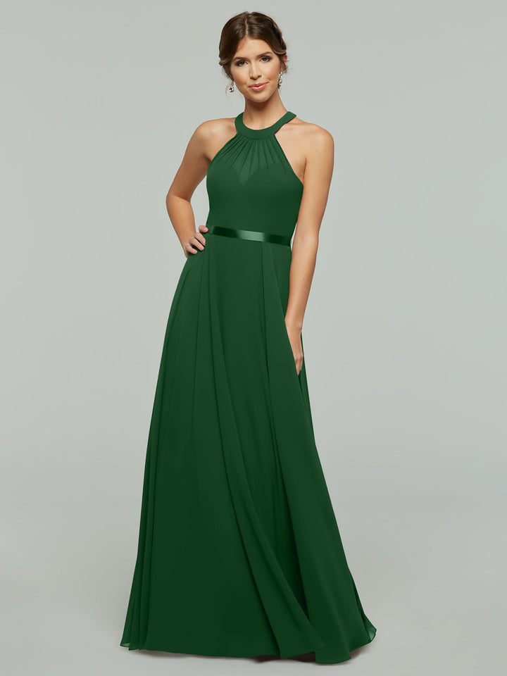 A woman poses against a light gray background, smiling slightly with one hand on her hip, wearing Avery Austin style 80038: a sleeveless, floor-length dark green A-line chiffon dress with a halter neckline and pleated skirt.