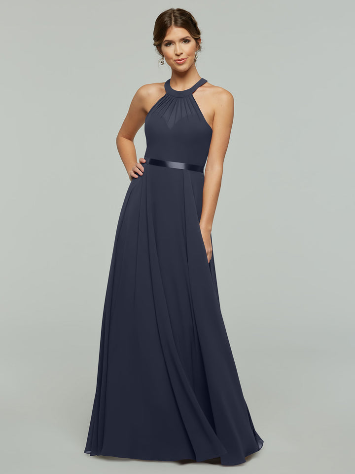 A woman models Avery Austin’s 80038, a navy blue sleeveless A-line chiffon gown with a halter neckline and fitted waist, standing before a plain light gray background.