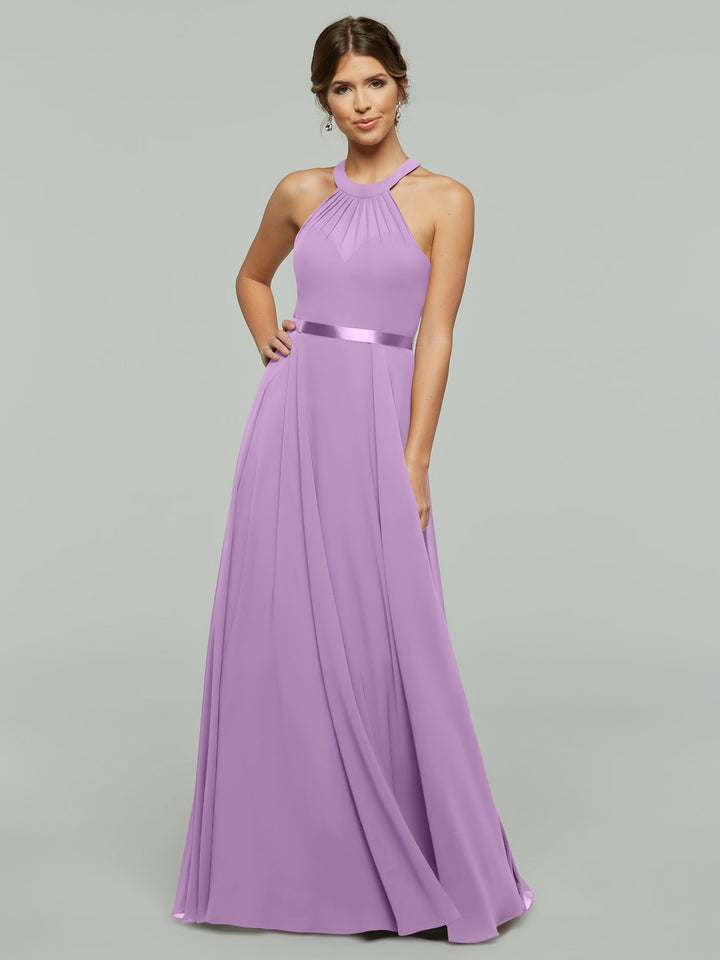 A woman in Avery Austin’s 80038 A-Line chiffon dress with a halter neckline and satin waistband stands against a light gray background, her hair up and smiling with one hand on her hip.