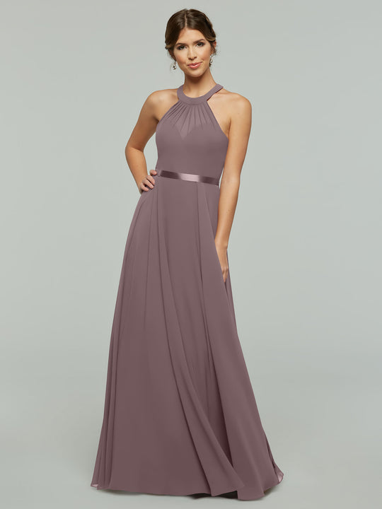 A woman wears Avery Austin’s 80038, an A-line mauve chiffon gown with a halter neckline and satin waistband, standing against a plain light gray background.