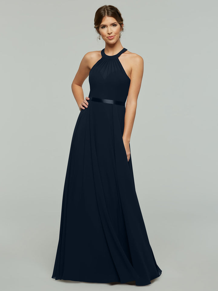 A woman models Avery Austin’s 80038: a sleeveless, floor-length dark navy blue A-line chiffon dress with a halter neckline, satin waistband, and pleated skirt. She poses against a plain light gray background, smiling slightly with one hand on her hip.