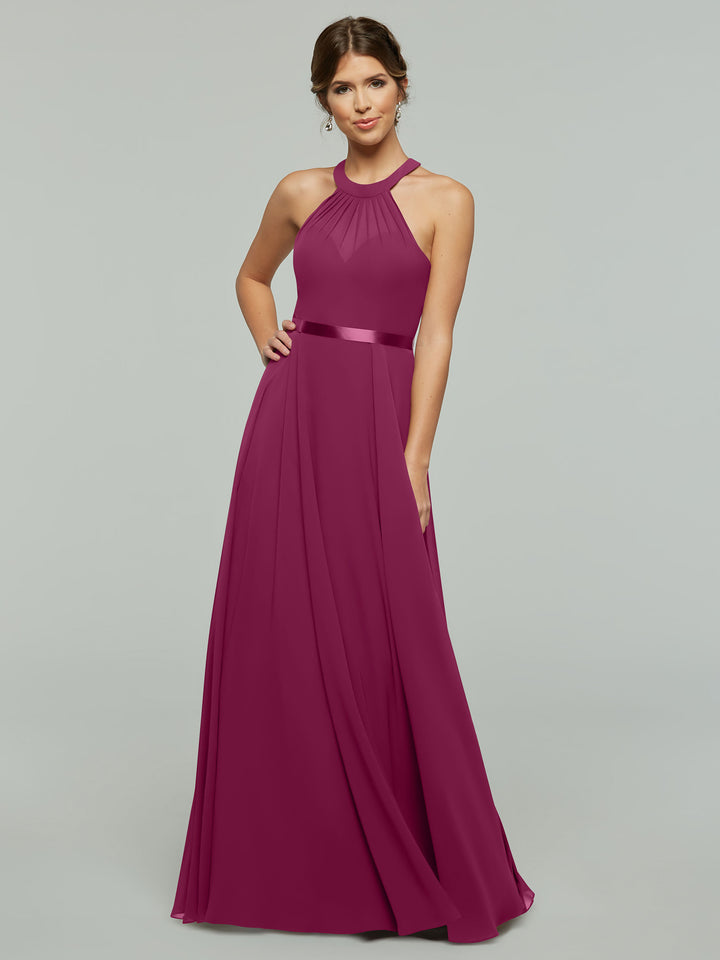 A woman poses against a plain background wearing Avery Austin style 80038, an A-line chiffon magenta dress with a halter neckline and satin waistband. Her hair is styled back, and she smiles softly with one hand on her hip.