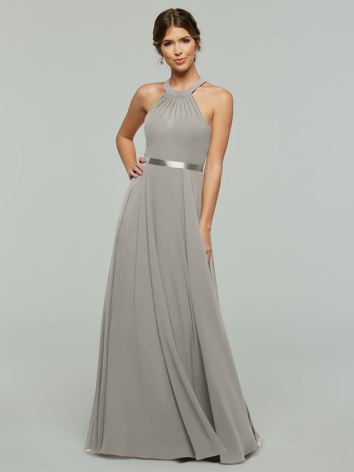 A woman models Avery Austin style 80038, an A-Line chiffon dress with a pleated skirt, halter neckline, and satin waistband, against a plain light gray background.