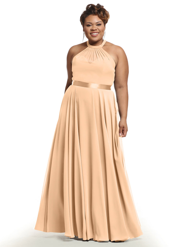A woman smiles in Avery Austin dress 80038, a blush A-line chiffon gown with halter neckline, pleated skirt, and satin waistband, standing before a plain white background.
