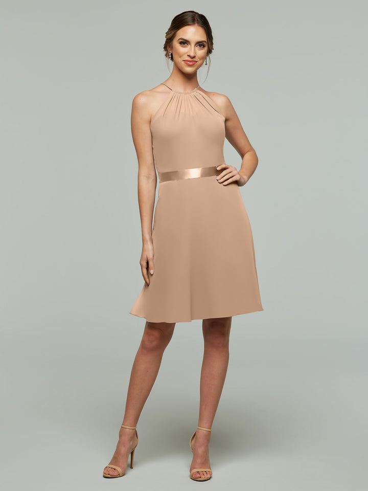 A model poses confidently with one hand on her hip, wearing Avery Austin’s 80042: a sleeveless beige dress featuring an A-line chiffon skirt and halter neckline, set against a plain light grey background.