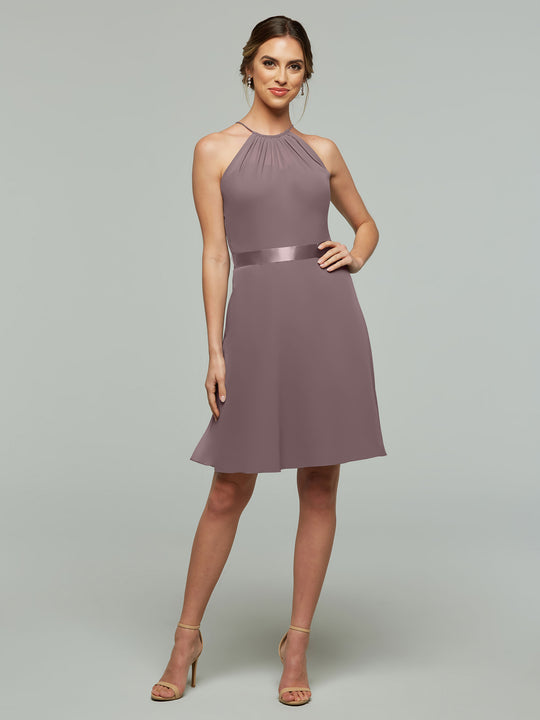 A woman models Avery Austin 80042, a sleeveless mauve halter dress with a keyhole back, A-line chiffon skirt, and satin waistband. She smiles in beige heels against a plain light gray backdrop.