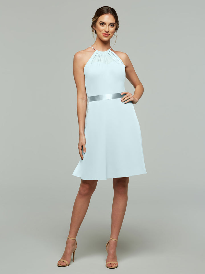 A woman in Avery Austin’s 80042: a sleeveless, light blue knee-length dress with an A-line chiffon skirt and satin waistband, paired with beige heels, stands smiling against a plain gray background.
