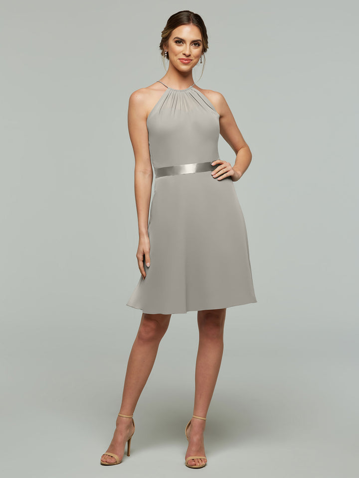 A woman models Avery Austin’s 80042 dress—a sleeveless, knee-length design with a halter neckline, keyhole back, satin waistband, and A-line chiffon skirt—standing against a plain light gray background with her hair styled up and open-toe heels.
