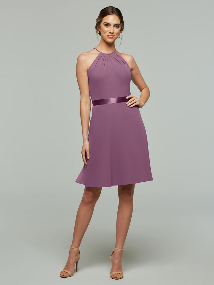 A woman wears Avery Austin style 80042, a sleeveless mauve halter dress with keyhole back, A-line chiffon skirt, and shiny waistband. She has light brown hair styled up, nude heels, and stands against a light gray background.