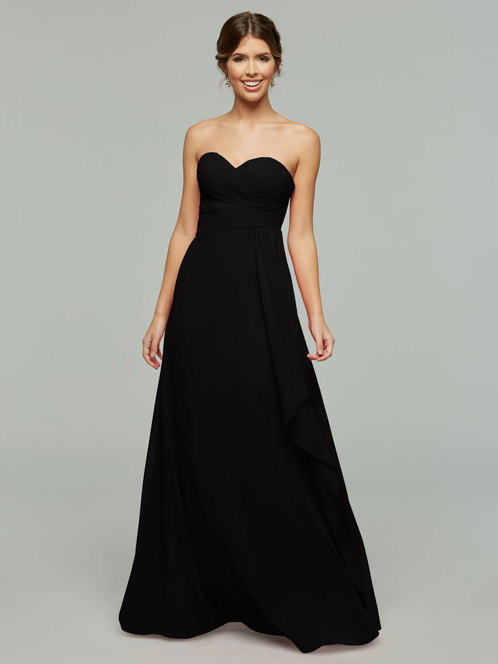 A woman smiles in the Avery Austin 80046, a timeless floor-length black gown with elegant draping and a sweetheart neckline, set against a plain light gray background.