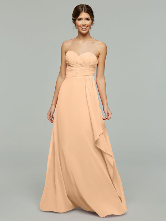 A woman wearing Avery Austin style 80046, a timeless strapless chiffon dress in peach with elegant draping and a ruched bodice, stands against a plain gray background, smiling with her hands by her sides.