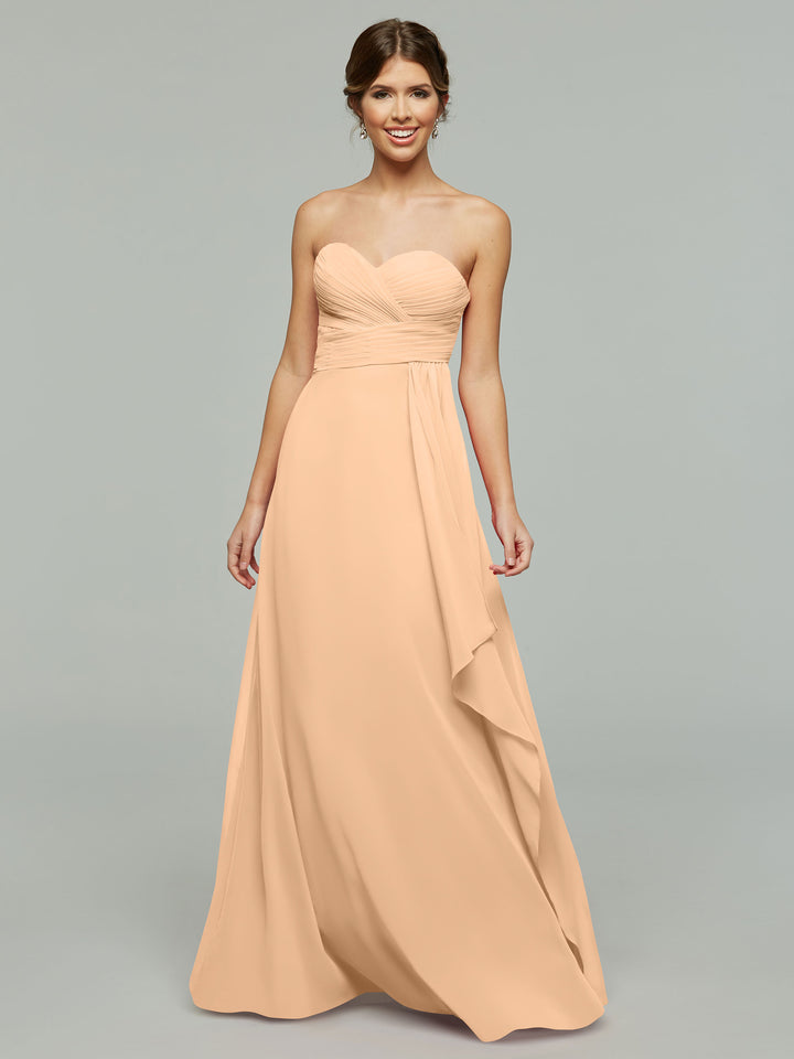 A woman wearing Avery Austin style 80046, a timeless strapless chiffon dress in peach with elegant draping and a ruched bodice, stands against a plain gray background, smiling with her hands by her sides.
