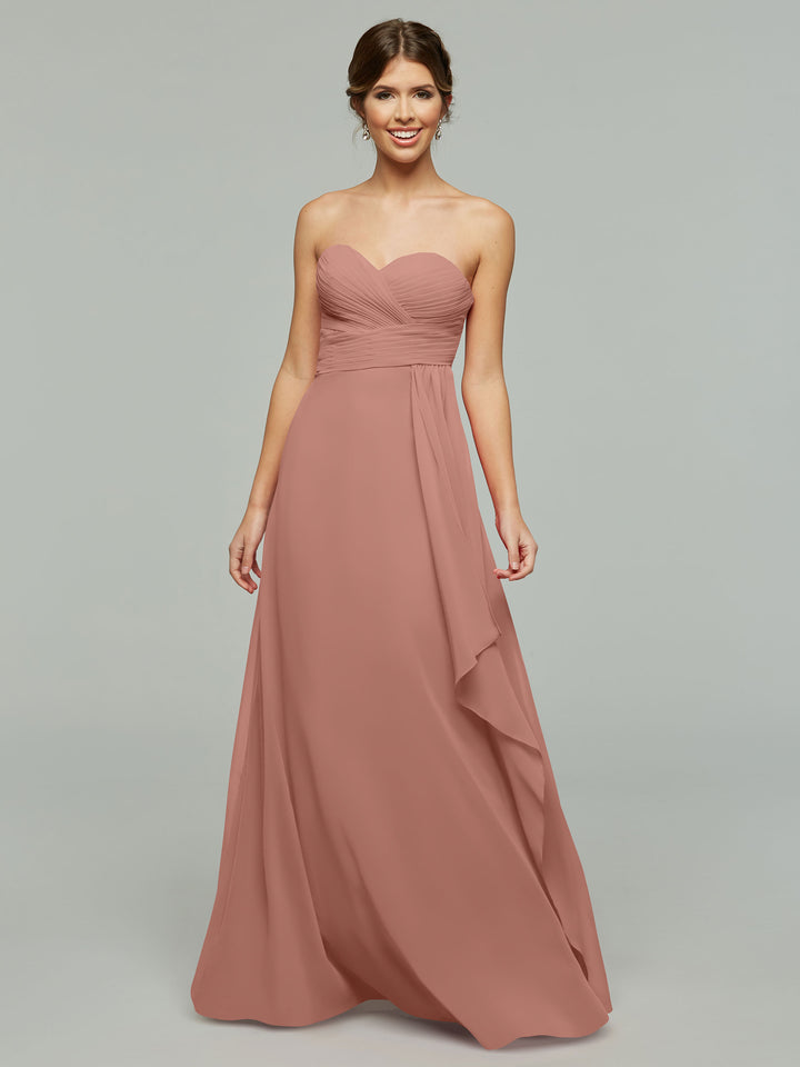 A woman in Avery Austin’s 80046, a strapless dusty rose chiffon dress with elegant draping and a ruched bodice, smiles against a plain light gray background.