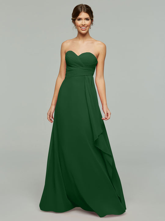 A woman wearing Avery Austin's 80046, a timeless dark green strapless chiffon dress with a fitted bodice and flowing skirt, stands and smiles against a plain light gray background.