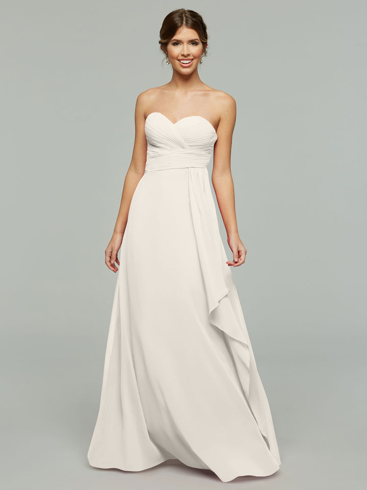 A woman smiles in the Avery Austin 80046, a timeless strapless chiffon gown with a sweetheart neckline and ruched bodice, its elegant draping flowing to the floor as she poses before a plain light gray background.