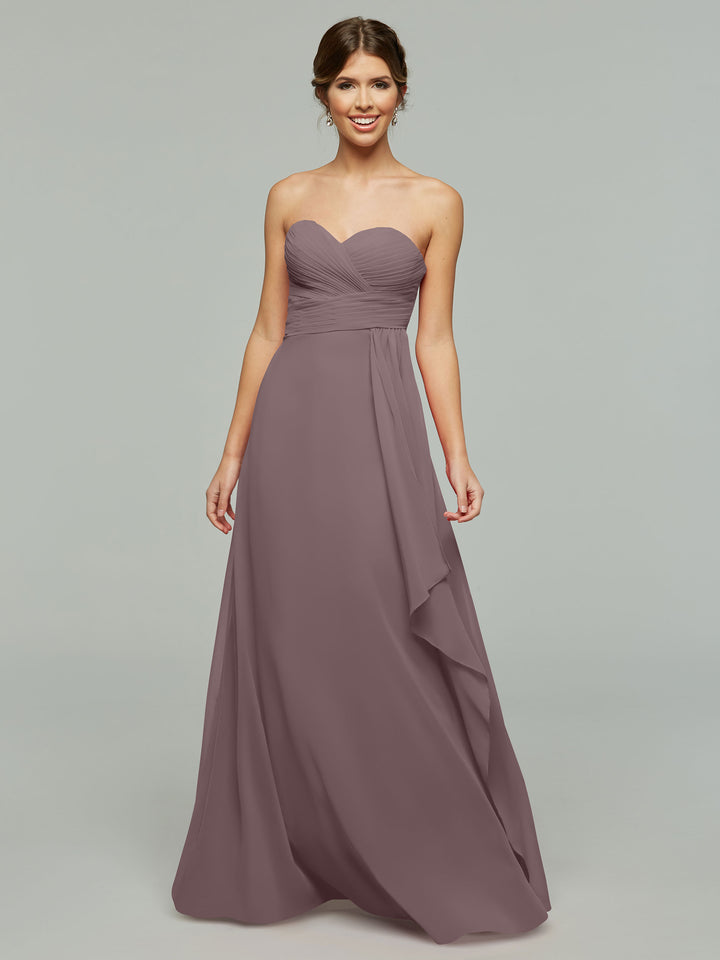 A woman smiles in Avery Austin’s 80046, a timeless strapless mauve chiffon dress with a sweetheart neckline, elegant draped waist, and floor-length skirt, set against a light gray background.