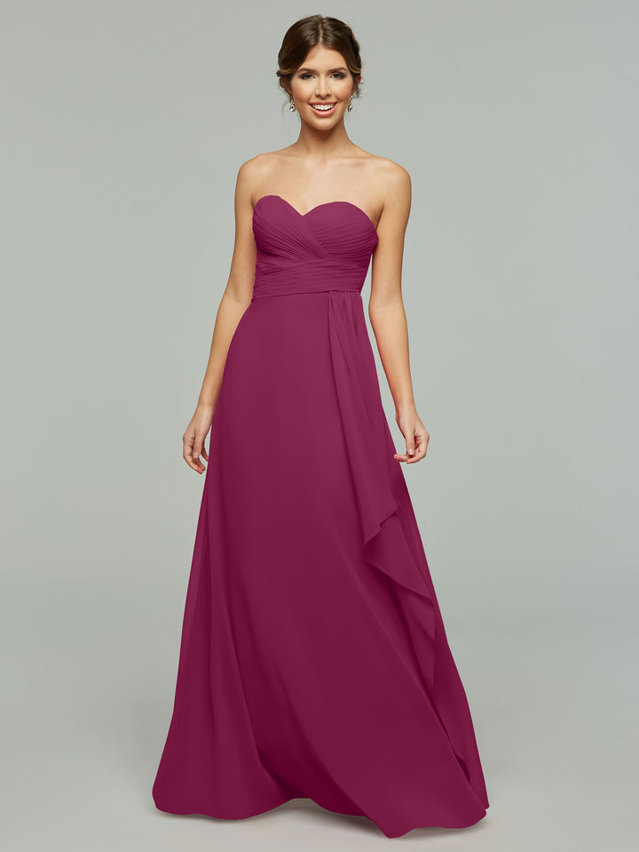 A woman in the Avery Austin 80046, a timeless strapless chiffon dress with a sweetheart neckline and elegant draping, stands smiling against a plain light gray background.